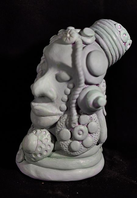 Face with thick lips, large nose and elaborate headress in coils and flower shapes sculpted in polymer clay. 
